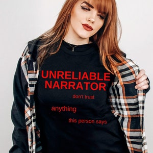 Puede incluir: Una camiseta negra con el texto "UNRELIABLE NARRATOR don't trust anything this person says" en letras rojas. La persona lleva una camisa de cuadros sobre la camiseta.