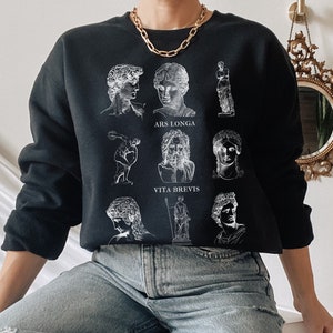 Peut inclure: Sweat-shirt noir avec des illustrations blanches de sculptures classiques et les phrases "ARS LONGA" et "VITA BREVIS". Le sweat-shirt a un col rond et des manches longues. Le modèle porte un jean clair déchiré et un collier chaîne doré.