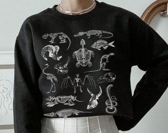 Op de afbeelding: Zwart gecropped sweatshirt met een witte print van verschillende dieren skeletten. De print bevat een schildpad, een vogel, een vis, een slang en andere wezens.