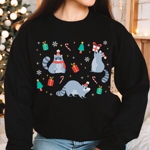 May include: Black sweatshirt with a Christmas design featuring raccoons wearing Santa hats and holding gifts. The design also includes candy canes, snowflakes, and Christmas trees.