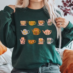 May include: A dark green sweatshirt with a floral teacup and teapot design. The design features a variety of teacups and teapots with floral patterns in shades of pink, yellow, and orange.