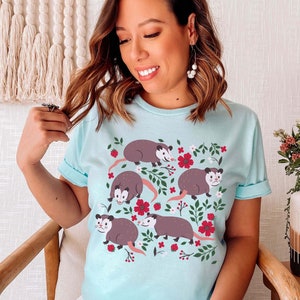May include: A light blue t-shirt with a floral pattern and multiple illustrations of opossums. The opossums are brown and gray with white accents. The flowers are red and white.