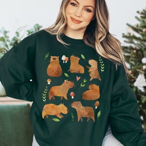 May include: A dark green sweatshirt with a pattern of brown capybaras and mushrooms. The capybaras are in various poses, including sitting, standing, and lying down. The mushrooms are red and white.