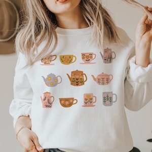 May include: White sweatshirt with a floral pattern of teapots and teacups. The design features a variety of colors, including pink, yellow, blue, and green.