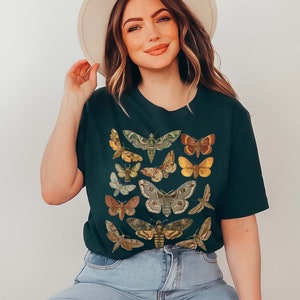 May include: A dark green t-shirt with a print of various butterflies and moths in shades of brown, yellow, and green. The shirt has a crew neck and short sleeves.