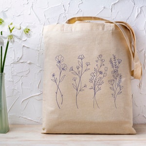 May include: A beige canvas tote bag with a design of five different floral sprigs in blue ink. The bag has two long straps.