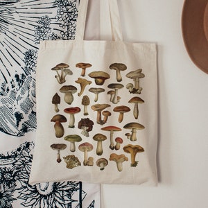 May include: A white canvas tote bag with a design of various mushrooms in shades of brown, red, and white. The mushrooms are arranged in a grid pattern on the bag.