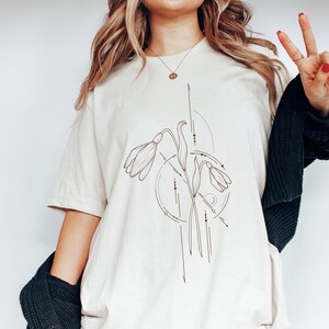 May include: A white t-shirt with a graphic of a stylized flower design in brown. The design features a flower with three petals and geometric lines.