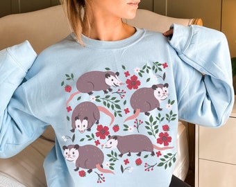 May include: A light blue sweatshirt featuring a graphic print of five opossums and flowers. The opossums are brown and grey with white accents. The flowers are red, pink, and white.