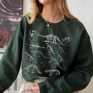 May include: A dark green sweatshirt with a white skeleton dinosaur print. The sweatshirt has a crew neck and long sleeves.