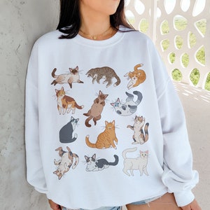 Op de afbeelding: Witte trui met een grafische print van verschillende katten in verschillende poses. De katten zijn in tinten bruin, zwart, wit en grijs.