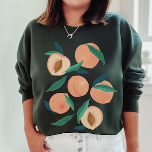 K&ouml;nnte beinhalten: Ein dunkelgr&uuml;ner Pullover mit einem Grafikdruck von Pfirsichen und Bl&auml;ttern. Der Pullover hat einen Rundhalsausschnitt und lange &Auml;rmel.