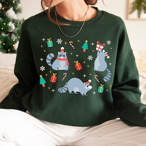 May include: A dark green sweatshirt with a festive Christmas design featuring raccoons wearing Santa hats and holding gifts. The design includes snowflakes, candy canes, and Christmas trees.
