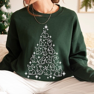 Pode incluir: Uma camisa de frio verde escura com um contorno branco de uma árvore de Natal feita de coelhos. A árvore é decorada com estrelas brancas.