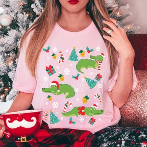 以下が含まれることがあります： サンタの帽子とマフラーをつけた緑色のワニが、クリスマスツリー、キャンディーケーン、雪片、ベルに囲まれているクリスマスをテーマにしたデザインのピンクのTシャツです。