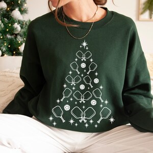 K&ouml;nnte beinhalten: Ein dunkelgr&uuml;ner Pullover mit einem wei&szlig;en Design eines Weihnachtsbaums aus Pickleball-Schl&auml;gern und B&auml;llen.