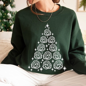 Op de afbeelding: Een donkergroen sweatshirt met een witte omtrek van een kerstboom die is gemaakt van hondenkoppen. De boom is versierd met witte sterren.