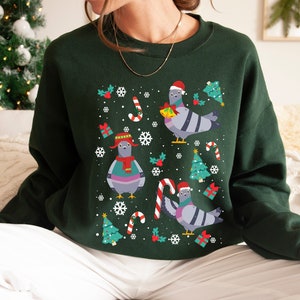May include: A green Christmas jumper with a festive design featuring cartoon pigeons wearing Santa hats and holding gifts. The jumper has a crew neck and long sleeves.