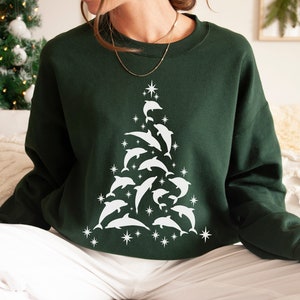 Puede incluir: Una sudadera verde oscuro con un diseño blanco de un árbol de Navidad hecho de delfines. El árbol está decorado con estrellas blancas.