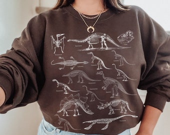 Op de afbeelding: Een bruin sweatshirt met een witte print van dinosaurus skeletten. Het sweatshirt heeft een ronde hals en lange mouwen. Het model draagt het sweatshirt met een blauwe denim short.