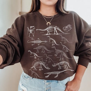 May include: A brown sweatshirt with a white print of dinosaur skeletons. The sweatshirt has a crew neck and long sleeves. The model is wearing the sweatshirt with blue denim shorts.