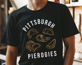 May include: Black t-shirt with the words "Pittsburgh Pierogies" in white, with an illustration of pierogies in gold. The shirt is short-sleeved and has a crew neck. The design is centered on the front of the shirt.
