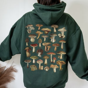 May include: Dark green hoodie featuring a detailed illustration of various mushrooms in shades of brown, red, and green. The design is printed on the back of the hoodie. The hoodie has a hood and long sleeves.