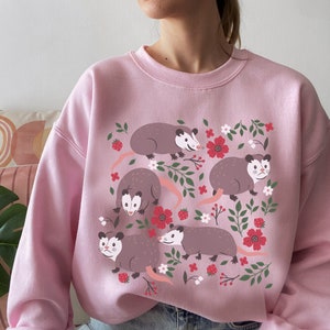 May include: Pink sweatshirt with a graphic print of four opossums in a floral design. The opossums are brown and gray with white accents. The flowers are red, pink, and white.