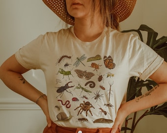 May include: A white t-shirt with a colorful insect print. The shirt features a variety of insects, including butterflies, beetles, spiders, and caterpillars. The shirt is being worn by a person with a straw hat.