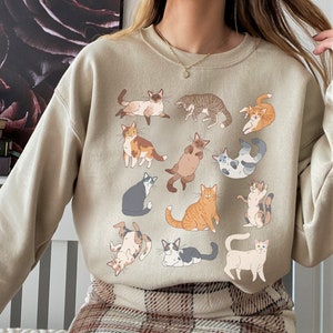 May include: A beige sweatshirt with a graphic print of various cats in different poses. The cats are all in shades of brown, white, and gray.