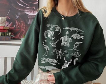 Op de afbeelding: Een donkergroen sweatshirt met een witte print van verschillende dieren skeletten. De print bevat een schildpad, een vis, een vleermuis, een vogel en andere wezens.