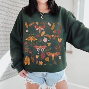 Puede incluir: Una sudadera verde oscuro con un patr&oacute;n de polillas, cristales y hojas. Las polillas son de varios tonos de marr&oacute;n, naranja y amarillo. Los cristales son de tonos rosa, morado y azul. Las hojas son de tonos verde, marr&oacute;n y naranja.