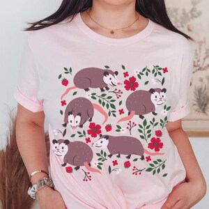 May include: A pink t-shirt with a graphic print of five opossums surrounded by red flowers and green leaves.