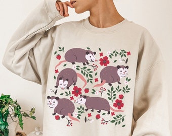 May include: A beige sweatshirt featuring a graphic print of several opossums surrounded by red flowers and green leaves.