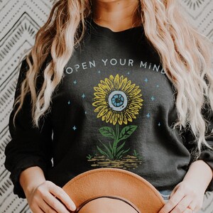 May include: Black sweatshirt with a sunflower graphic and the text "Open Your Mind". The sunflower has an eye in the center.