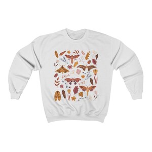 Puede incluir: Sudadera blanca con un patr&oacute;n colorido de polillas, cristales y flores. Las polillas son marrones, naranjas y rosas con patrones intrincados. Los cristales son transparentes y brillantes. Las flores son amarillas, rosas y naranjas.