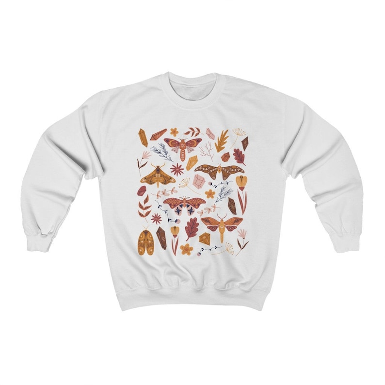 Puede incluir: Sudadera blanca con un patr&oacute;n colorido de polillas, cristales y flores. Las polillas son marrones, naranjas y rosas con patrones intrincados. Los cristales son transparentes y brillantes. Las flores son amarillas, rosas y naranjas.