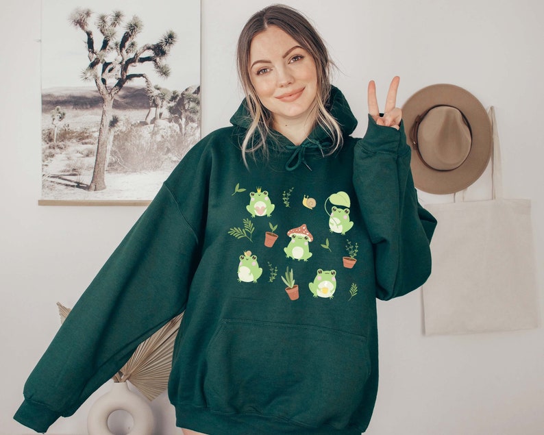 May include: A dark green hooded sweatshirt with a graphic print of frogs, mushrooms, and plants. The sweatshirt has a drawstring hood and a kangaroo pocket.