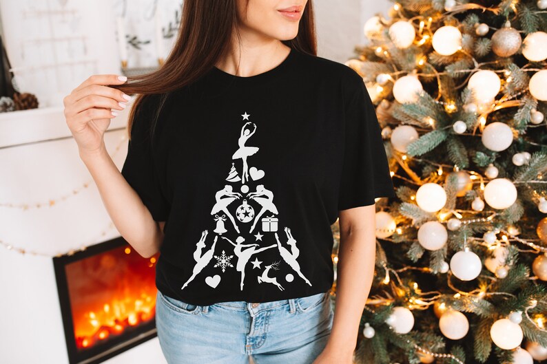 May include: A black t-shirt with a white design of a Christmas tree made up of dancers and Christmas ornaments. The tree is topped with a star.