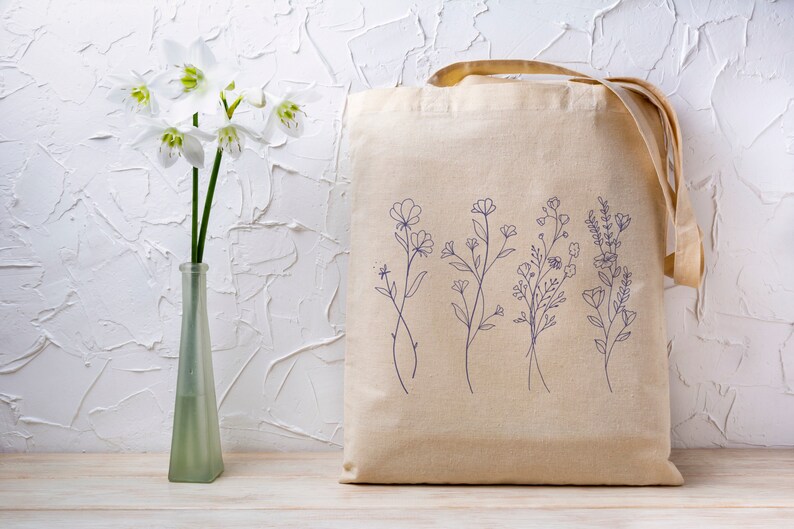 May include: A beige canvas tote bag with a design of five different floral sprigs in blue ink. The bag has two long straps.