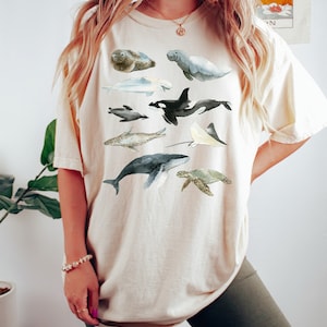 May include: A beige t-shirt with a watercolor illustration of various sea creatures, including a whale, a dolphin, a sea turtle, a stingray, and a seal.