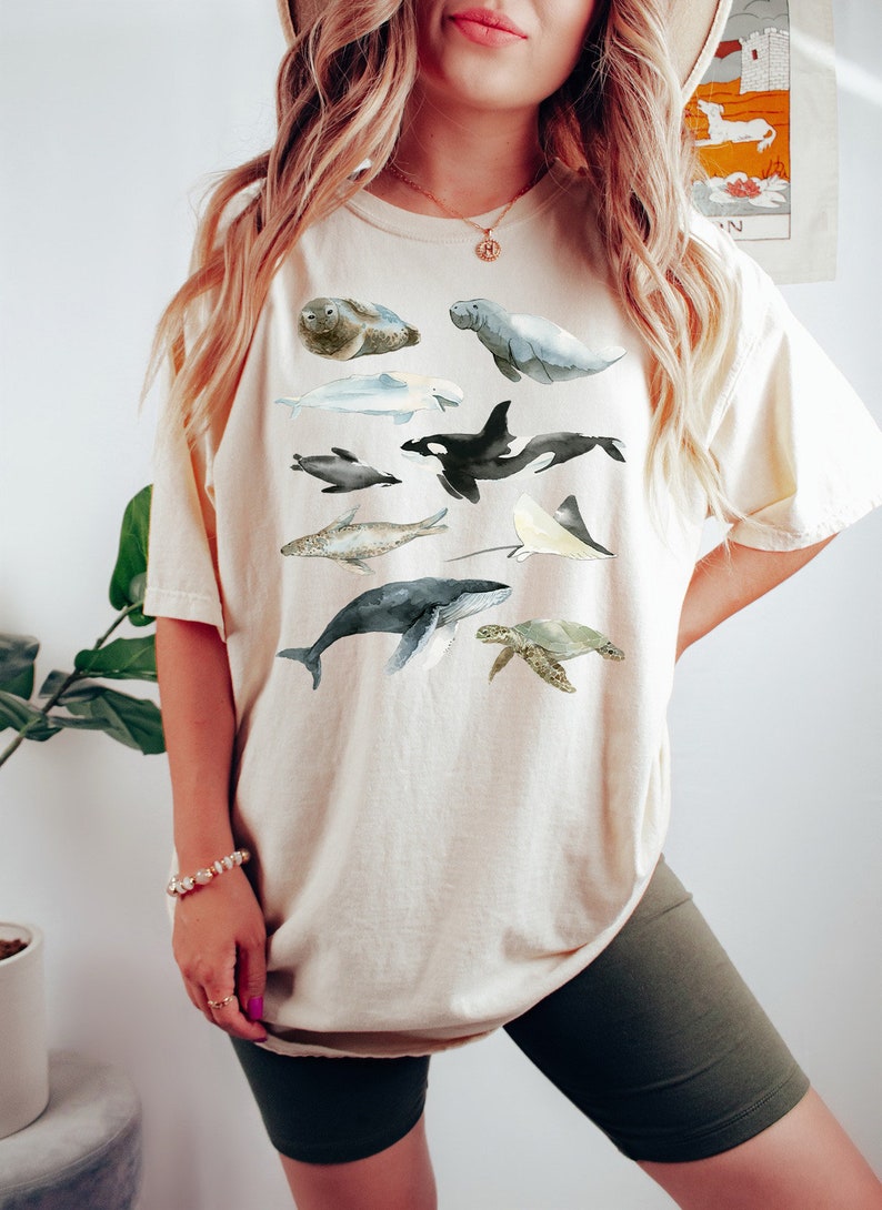 May include: A beige t-shirt with a watercolor illustration of various sea creatures, including a whale, a dolphin, a sea turtle, a stingray, and a seal.