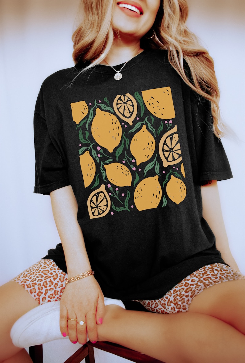 Puede incluir: Camiseta negra con estampado de limones. Los limones son amarillos con hojas verdes y flores rosas.
