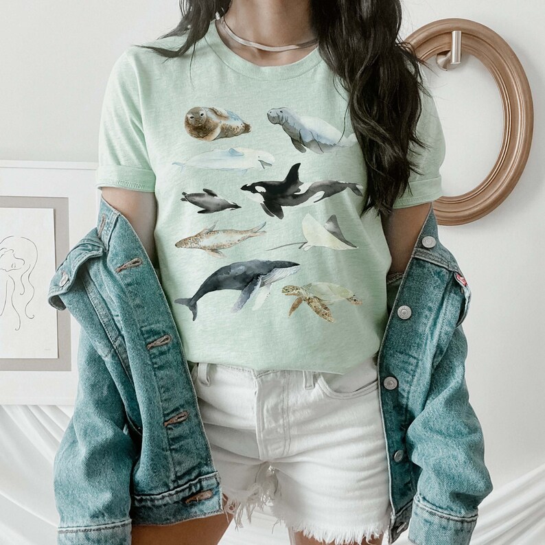 K&ouml;nnte beinhalten: Ein hellgr&uuml;nes T-Shirt mit einem Aquarell-Design aus verschiedenen Meerestieren, darunter ein Wal, ein Delfin, eine Meeresschildkr&ouml;te, ein Rochen und eine Robbe. Das Shirt wird von einer Person mit einer Jeansjacke getragen.