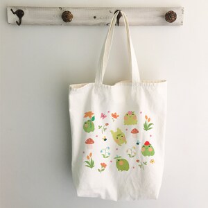 May include: A white canvas tote bag with a design of green frogs, flowers, and mushrooms. The frogs are wearing flowers on their heads and some have a red hat.