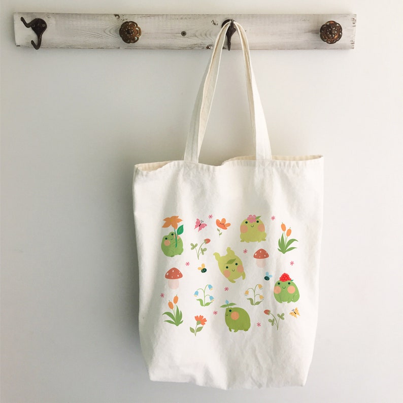 May include: A white canvas tote bag with a design of green frogs, flowers, and mushrooms. The frogs are wearing flowers on their heads and some have a red hat.