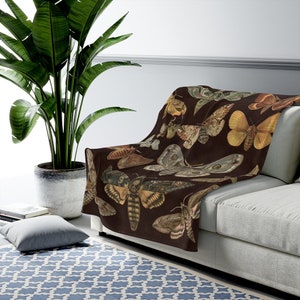 May include: A brown throw blanket with a repeating pattern of colorful moths and butterflies. The blanket is draped over a light gray couch.