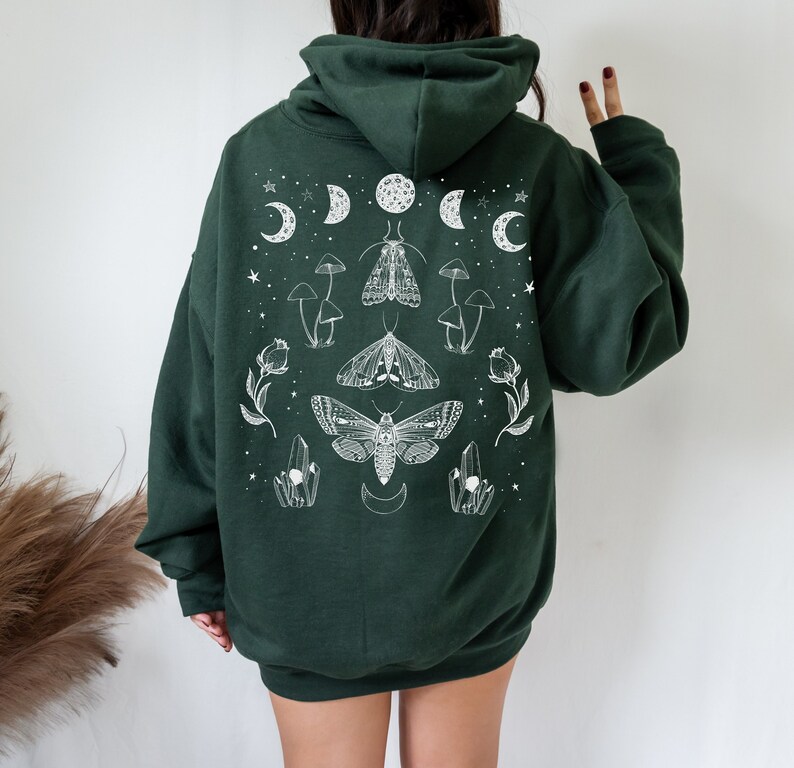 Pu&ograve; includere: Una felpa con cappuccio verde scuro con una stampa grafica bianca di falene, funghi, cristalli e lune crescenti.