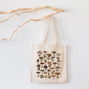 May include: A white canvas tote bag with a design of various mushrooms in shades of brown, red, and white. The mushrooms are arranged in a grid pattern on the bag.