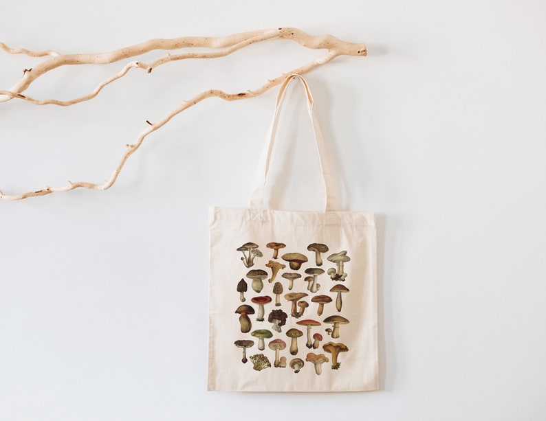 May include: A white canvas tote bag with a design of various mushrooms in shades of brown, red, and white. The mushrooms are arranged in a grid pattern on the bag.